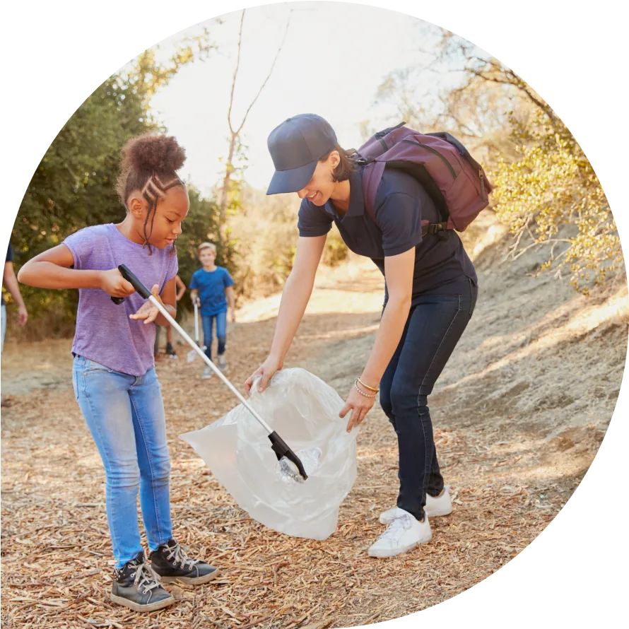 A woman with a maroon backpack helps a young girl put trash into a bag.