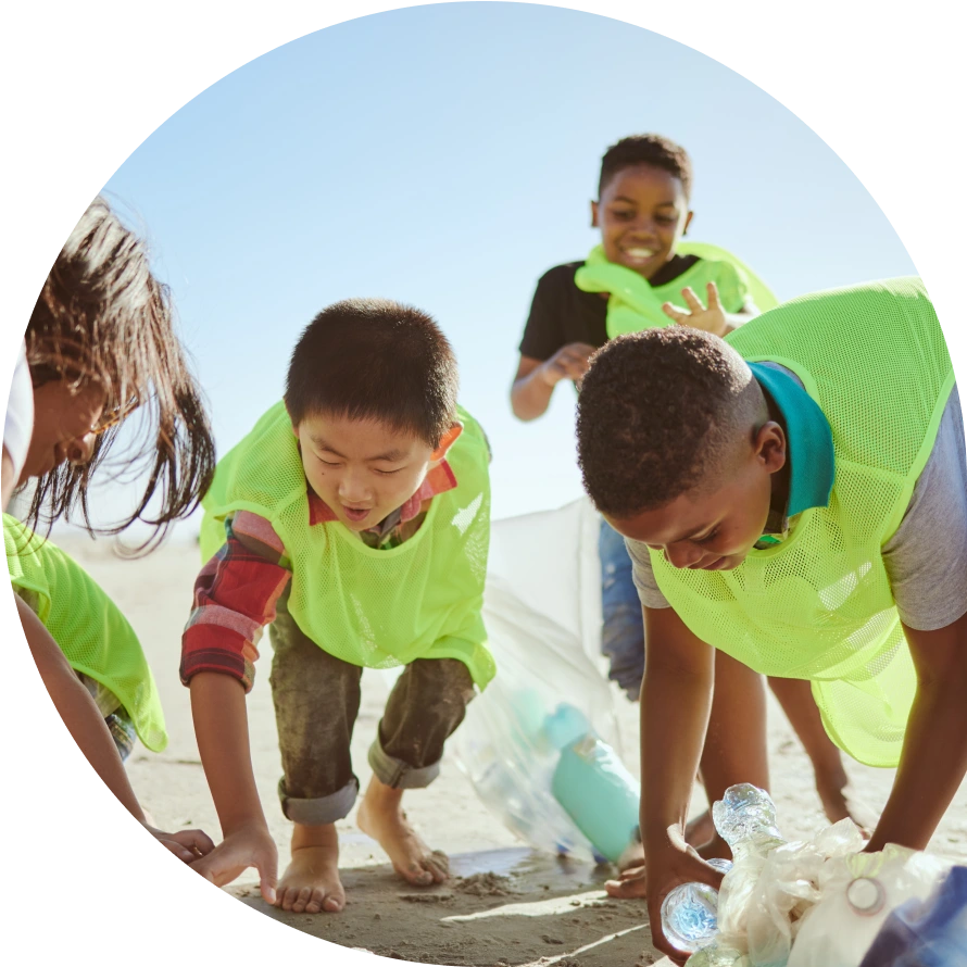 Four young children in yellow vests pick up trash on a sandy beach.